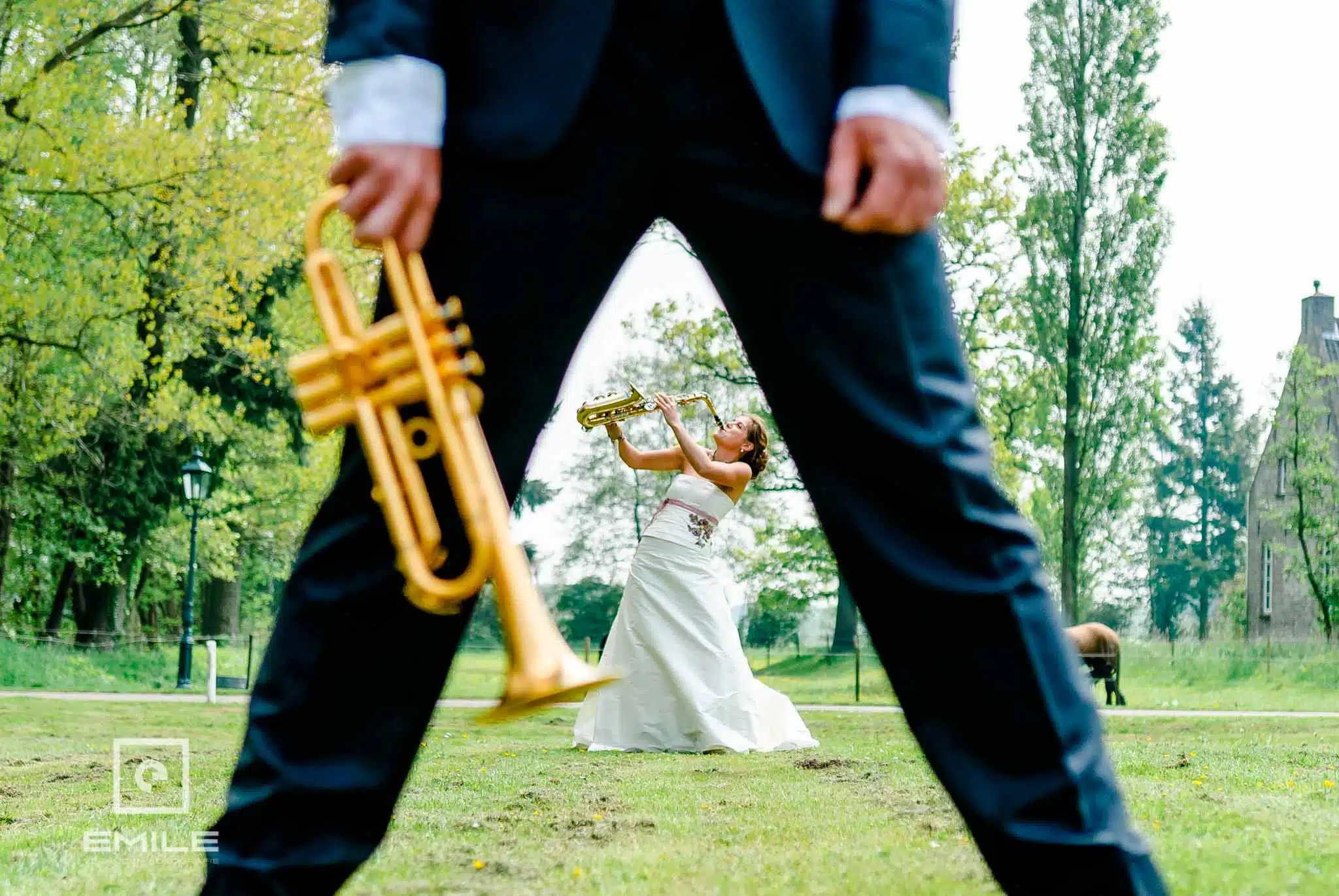 Op de foto is een bruidspaar te zien waarbij de bruid op een saxofoon speelt tussen de benen door van de bruidegom. Hij staat met zijn benen iets uit elkaar en houd zijn trompet vast en de bruid speelt op de saxofoon. De bruid heeft een intense blik in haar ogen en speelt met passie. De foto is genomen op een mooie, zonnige dag in de wei bij Kasteel Daelenbroeck te Herkenbosch