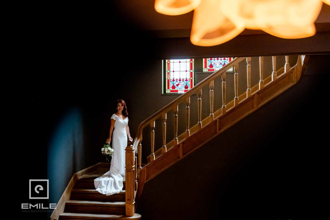 Op de foto is een prachtige bruid te zien die op de trap van Kasteel Hoogenweerth Maastricht staat. De trap om haar heen is indrukwekkend, met hoge muren, glas-in-lood en een hoog plafond. Er is een kristallenlamp aan het plafond bevestigd die prachtig warm licht geeft. Trouwfotograaf Limburg