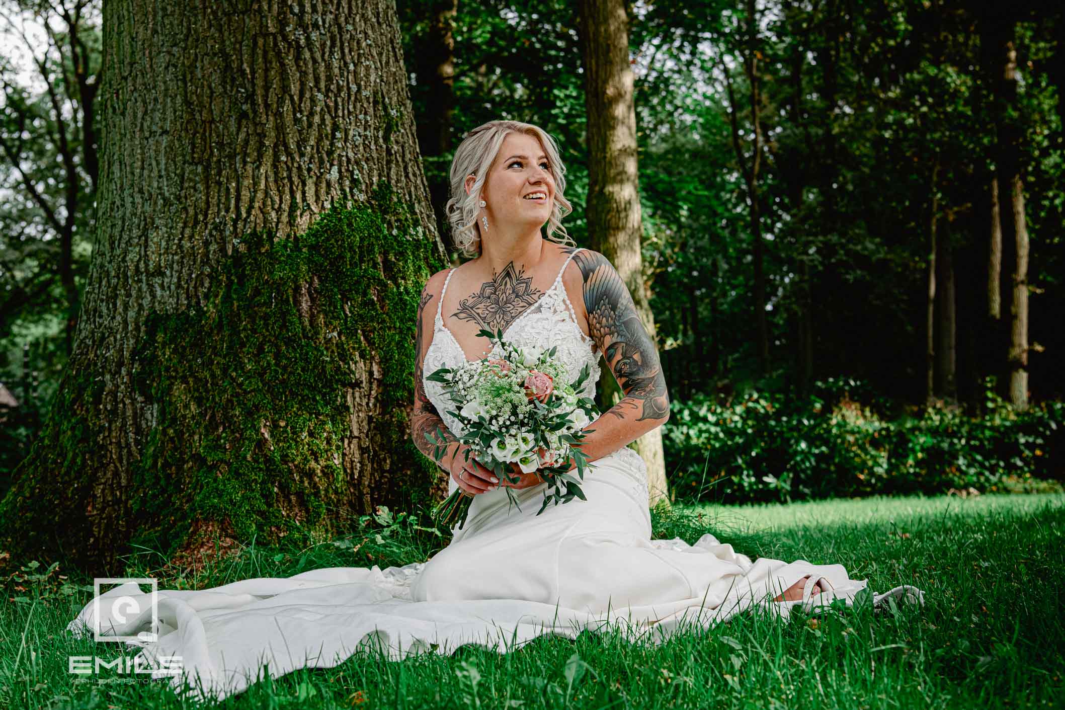 Bruid met veel tattoos zit bij boom op de grond in de Brunssumerheide